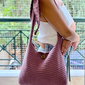 May include: A mauve-colored, crocheted shoulder bag with a large wooden button closure. The bag has a wide strap and a rounded shape. The background shows a green, outdoor setting.