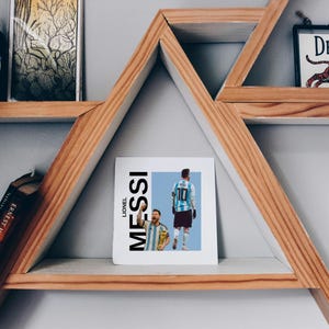 Peut inclure: Impression carrée avec Lionel Messi en uniforme de foot, le nom "MESSI" en gras et vertical. L'impression est sur une étagère en bois, avec un réveil noir, une image encadrée et un livre.