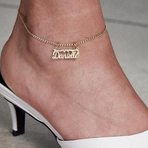 Può includere: Una cavigliera color oro con il nome "Danielle" in corsivo. La cavigliera presenta una catena delicata e piccoli cuori. La persona indossa scarpe bianche senza lacci con un piccolo tacco.