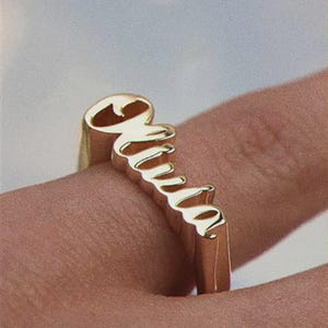 May include: A gold-colored ring spelling out the name "Mia" in a cursive font. The ring is worn on a finger, showcasing the personalized design. The ring's letters are raised, creating a 3D effect.