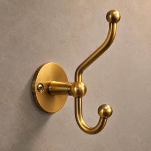 Puede incluir: Un gancho doble para pared con acabado en latón cepillado. El herraje presenta una placa base circular con un tornillo visible, un poste horizontal central y dos brazos curvos que terminan en pomos esféricos para colgar prendas.