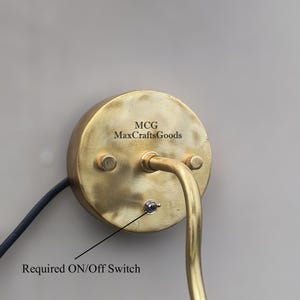 May include: A brass-colored wall sconce with a curved arm and a round backplate. The backplate has the text "MCG MaxCraftsGoods" and two small screws. It features an on/off switch and a black power cord.