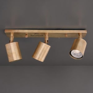 May include: A brass-coloured, three-light ceiling spotlight fixture. The fixture has three adjustable cylindrical spotlights attached to a rectangular base. The text "MCG MaxCraftGoods" is printed on the base.