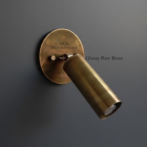 May include: A brass wall-mounted spotlight with a round backplate and a cylindrical light fixture. The fixture is angled and labeled "Glossy Raw Brass". The backplate has the text "MCG MaxCraftsGoods".