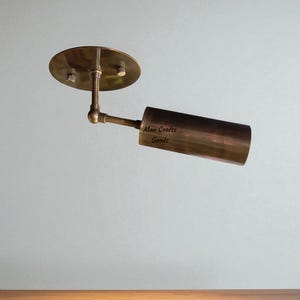 Puede incluir: Un foco de techo ajustable de color bronce. Cuenta con una base circular con dos tornillos, un brazo giratorio y una luminaria cilíndrica. El texto "Max Crafts Goods" está impreso en la luminaria.