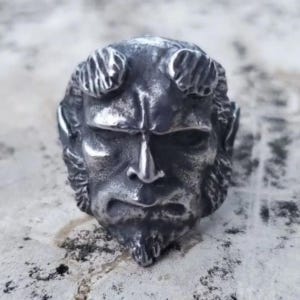 May include: A silver-toned ring featuring a detailed, sculpted face with horns, a goatee, and a stern expression. The ring has a textured, aged appearance, suggesting a vintage or gothic style. The design is a close-up of a character's face.