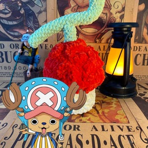 Op de afbeelding: Een gehaakte, rode pluche vrucht met een groene stengel, die lijkt op een duivelsvrucht uit de anime-serie One Piece. Een stripfiguur, Tony Tony Chopper, staat op de voorgrond. Rechts staat een kleine lantaarn.