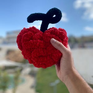 Op de afbeelding: Een handgemaakte, pluche rode kers met een donkergroene steel. De kers wordt tegen een blauwe luchtachtergrond gehouden. De kers is gemaakt van een zacht, getextureerd materiaal.