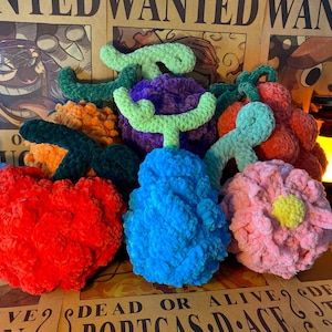 Puede incluir: Una colección de frutas y flores de ganchillo, coloridas y de felpa. El surtido incluye una manzana roja, una baya azul, una flor rosa, una fruta naranja, una fruta morada y uvas rojas, cada una con tallos verdes. Los artículos se exhiben en un póster vintage.