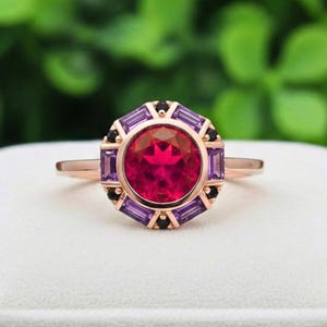 May include: Rose gold ring featuring a large, round, red gemstone at the center, surrounded by smaller rectangular purple and black stones. The ring has a delicate band and a detailed, geometric design around the main stone.