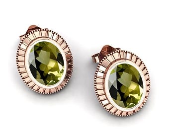 May include: A pair of rose gold stud earrings. Each earring features an oval, faceted, light green gemstone set within a decorative, circular frame. The frame has a textured, ridged design around the gemstone.