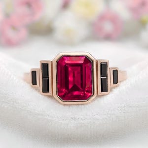 May include: Rose gold ring with a large, rectangular, ruby-red gemstone. The ring has a geometric design with black accents. The ring is set against a white background. The ring's design is Art Deco inspired.