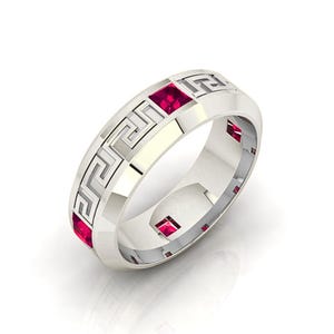 May include: A silver ring with a Greek key design and square, red gemstones. The ring has a polished finish and a classic, geometric pattern. The gemstones are evenly spaced around the band, adding a pop of color to the design.