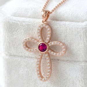 May include: Rose gold pendant necklace featuring a cross-shaped design. The pendant is adorned with a central, round, red gemstone and is embellished with small, clear gemstones. The necklace is displayed on a white, textured surface.
