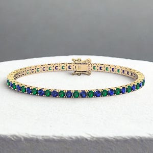 May include: A gold-toned tennis bracelet featuring alternating green and blue gemstones. The bracelet has a secure clasp and is displayed on a white surface. The gemstones are evenly spaced, creating a continuous line of color.