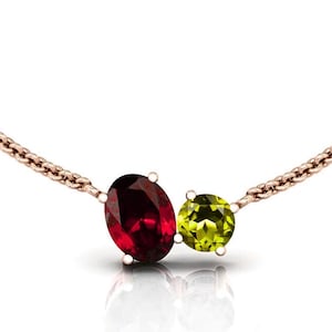 May include: A rose gold necklace featuring two gemstones: a large oval-cut red garnet and a smaller round-cut peridot. The gemstones are set close together, creating a unique pendant design. The chain is delicate and complements the gemstones.