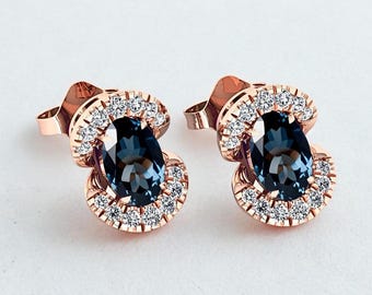 Könnte beinhalten: Ein Paar Ohrstecker aus Roségold. Jeder Ohrring hat einen ovalen, dunkelblauen Edelstein, der von einem Halo aus kleinen, klaren Edelsteinen umgeben ist. Die Ohrringe sind vor einem weißen Hintergrund platziert.