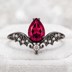 May include: A black metal ring featuring a central pear-shaped deep red gemstone. The band is designed with symmetrical bat wing motifs on each side, accented with small clear round stones along the wings and a single clear stone at the base.