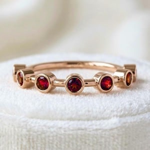 May include: A rose gold band ring featuring five round, bezel-set red gemstones arranged in a row. The metal has a polished finish, and each stone is securely held within a circular setting, creating a continuous, symmetrical design across the band.