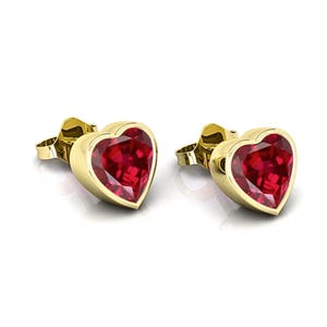 May include: A pair of heart-shaped stud earrings. Each earring features a vibrant red gemstone set in a gold-colored bezel. The earrings are displayed on a white background, highlighting their polished finish and romantic design.