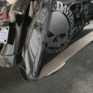 Puede incluir: Motocicleta Harley-Davidson negra con un gráfico de calavera en el lateral. La motocicleta tiene una placa de matrícula y un tubo de escape cromado. El logotipo de Harley-Davidson es visible.