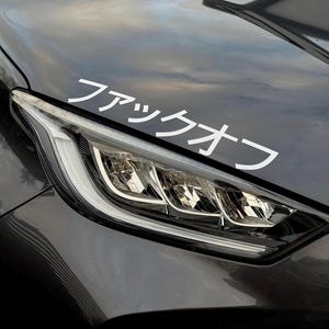 May include: A close-up of a dark gray car hood with a white decal. The decal features Japanese characters. The car's headlight is visible, with a bright, modern design. The car's paint reflects the sky.
