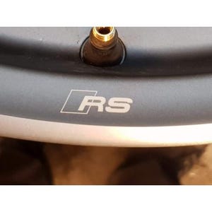 May include: Close-up of a section of a dark gray car wheel rim. The rim features the white letters "RS" inside a white rectangular outline. A gold-colored valve stem is visible at the top of the rim.