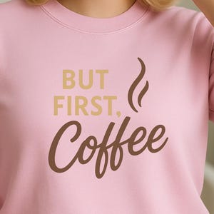 Könnte beinhalten: Hellrosa Sweatshirt mit dem Aufdruck "BUT FIRST, Coffee" in Gold und Braun. Das Design beinhaltet eine Kaffeebild-Grafik. Das Sweatshirt hat einen Rundhalsausschnitt.