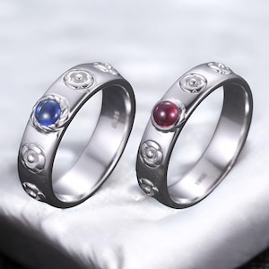 以下が含まれることがあります： 花柄と宝石のアクセントが付いた2つのシルバーリング。1つのリングには青い宝石、もう1つには赤い宝石が付いています。リングは白い表面に表示されています。