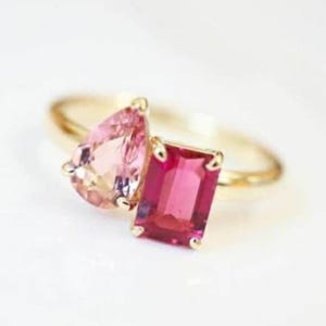 May include: A gold ring with two gemstones: a pear-shaped, light pink stone and a rectangular, deep pink stone. The ring's band is a simple, polished gold.