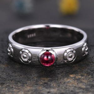 May include: A silver ring with a central, round, deep red gemstone. The band features a repeating floral pattern and is set against a dark background. The ring is a piece of jewelry.