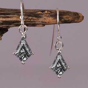 Puede incluir: Un par de pendientes colgantes de plata. Cada pendiente presenta una gema negra y gris en forma de diamante con detalles plateados. Los pendientes cuelgan de alambres de plata, mostrando un diseño moderno y elegante.