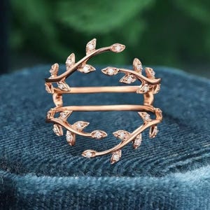 Può includere: Anello in oro rosa con un design a doppia fascia. L'anello presenta un delicato design a foglie e diamanti. Le foglie sono adornate da piccoli diamanti scintillanti. L'anello è posto su uno sfondo di velluto blu.