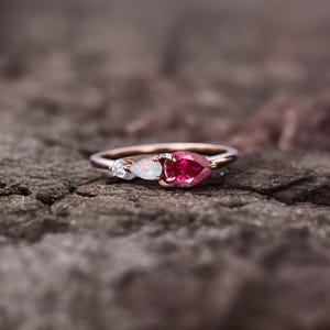 Peut inclure: Une bague en or rose ornée de trois pierres précieuses. Un rubis rouge vif en forme de poire est au centre, flanqué d'une opale blanche et d'un petit diamant. La bague est posée sur un fond texturé et neutre.