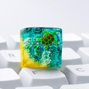 May include: A square resin mechanical keyboard keycap featuring a miniature green sea turtle with brown spots encased in a clear blue aquatic environment with yellow sand-like textures at the base, resting on white keyboard keys.