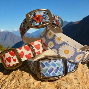 May include: A collection of patterned dog collars with silver-colored buckles. Designs include red hearts on white, blue and white floral, blue and white daisies, and blue brushstrokes. The collars are displayed on a rock with a mountain backdrop.