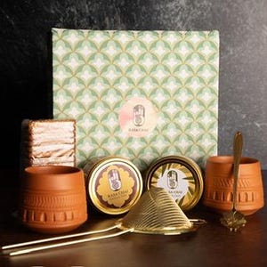 May include: A tea set featuring terracotta cups, a gold-colored tea strainer, and two tins of tea. The set includes a decorative box with a green and white geometric pattern and a logo. A small cake is also included.
