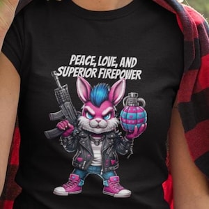 Può includere: Maglietta nera con un coniglio dei cartoni animati rosa e blu, con una giacca di pelle e che tiene in mano un mitra e una granata. Il testo "PEACE, LOVE, AND SUPERIOR FIREPOWER" è stampato sopra il coniglio.