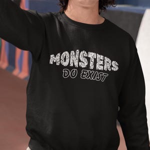May include: Black sweatshirt with the text "MONSTERS DO EXIST" in distressed white lettering. The sweatshirt is a crew neck style, and the text is centered on the chest.