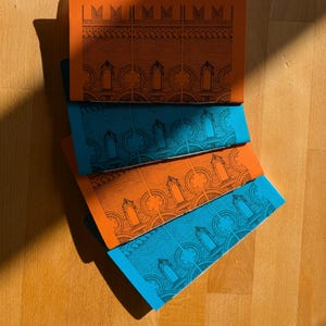 May include: A stack of orange and teal notebooks with architectural illustrations. The notebooks are arranged in a fan shape, revealing the patterned designs on the covers. The covers feature a repeating pattern of arched windows and decorative elements.
