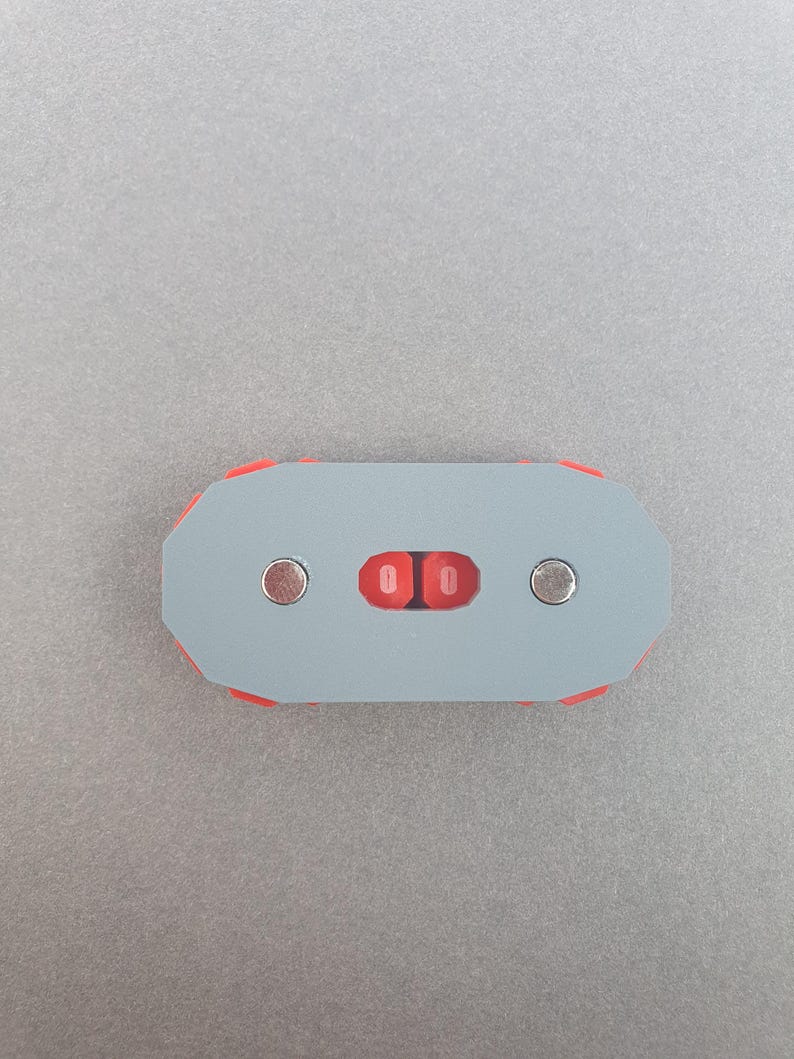Puede incluir: Un objeto rectangular gris con detalles rojos y dos imanes integrados. El centro presenta dos incrustaciones rojas de forma ovalada. El objeto est&aacute; dise&ntilde;ado con una forma geom&eacute;trica y una est&eacute;tica minimalista.