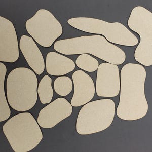 May include: A collection of irregularly shaped, light brown cardboard cutouts arranged on a gray surface. The cutouts vary in size and shape, resembling organic forms. The neutral color palette and varied forms suggest a craft or design project.