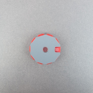 May include: A gray and red round object with a magnetic center and a small tab labeled "10". The object has a geometric design with a red outer edge and a gray center. The object is on a gray surface.