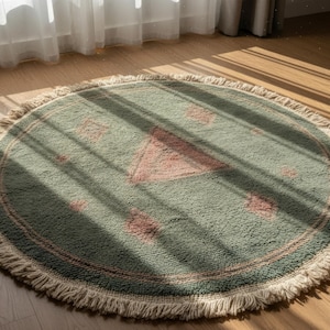 May include: A round, sage green rug with a pink geometric design and a cream-colored fringe. The rug is made of a soft, textured material and is placed on a wooden floor. The rug's diameter is approximately 4 feet.