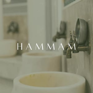 Op de afbeelding: Een spa-achtige setting met witte wastafels en antieke messing kranen. Het woord "HAMMAM" wordt weergegeven in een minimalistisch lettertype. De scène roept een gevoel van rust en traditioneel baden op.