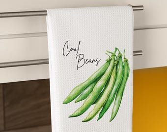 Op de afbeelding: Witte keukendoek met de tekst "Cool Beans" en een aquarel illustratie van sperziebonen. De handdoek hangt aan een zilveren stang in een keuken.