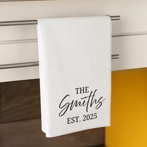 以下が含まれることがあります： 銀色のバーに掛けられた、テクスチャード加工のワッフル織りの白いキッチンタオル。「THE Smiths EST. 2025」の文字がスタイリッシュな筆記体で書かれており、パーソナライズされたギフトに最適です。
