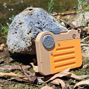 May include: A small, tan and orange camera-shaped object with a circular dial and rectangular body. The object is resting on a bed of leaves and twigs, with a large rock in the background. The camera has a retro design.