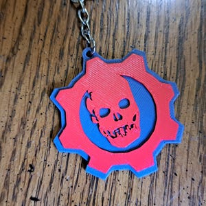 Puede incluir: Un llavero rojo y azul con un diseño de calavera dentro de una forma de engranaje. El llavero está impreso en 3D y sujeto a una cadena plateada. El diseño es una calavera estilizada. Probablemente sea de plástico.