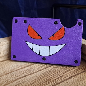 May include: A purple card holder featuring a cartoon face with orange eyes and a wide, white-toothed grin. The card holder has a cut-out section and is secured with black rivets. The design is reminiscent of a popular animated character.